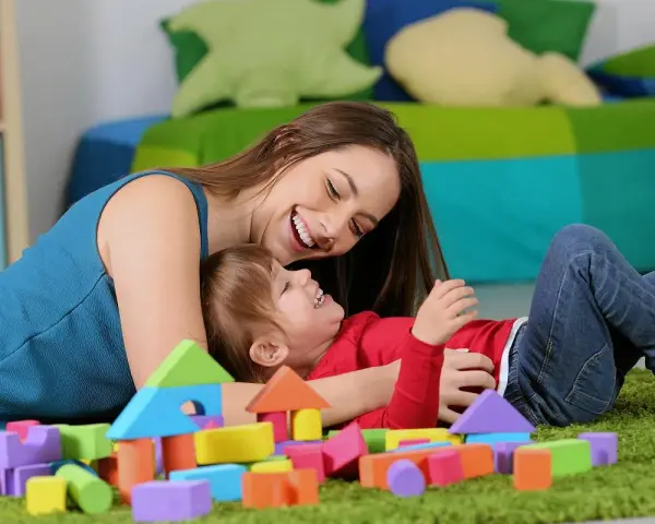 Uśmiechnięta opiekunka bawi się z dzieckiem wśród kolorowych klocków. Zastanawiasz się, ile kosztuje opieka nad dzieckiem za godzinę?