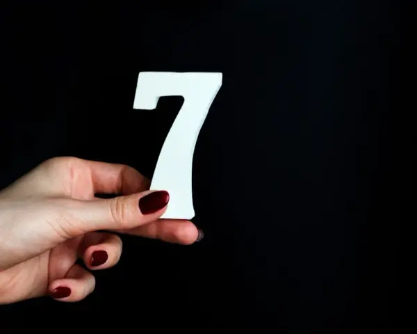 Dłoń z ciemnoczerwonym lakierem trzyma białą cyfrę 7. Może to symbolizować numerologiczną 7 mężczyzny, który jest tajemniczy i introspektywny.