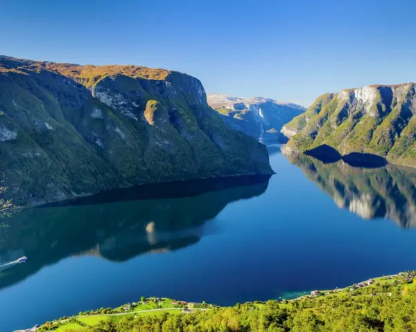 En båt färdas på en spegelblank fjord i Norge, omgiven av gröna bergssluttningar. En tågresa genom detta landskap vore en dröm.