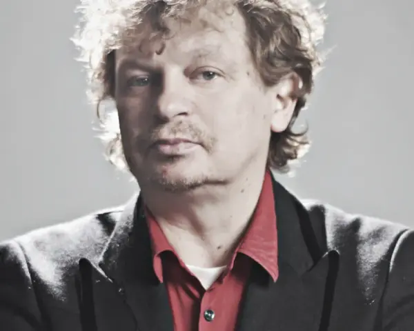 Marek Zieliński, aktor, w czarnej marynarce i czerwonej koszuli, z kręconymi włosami.