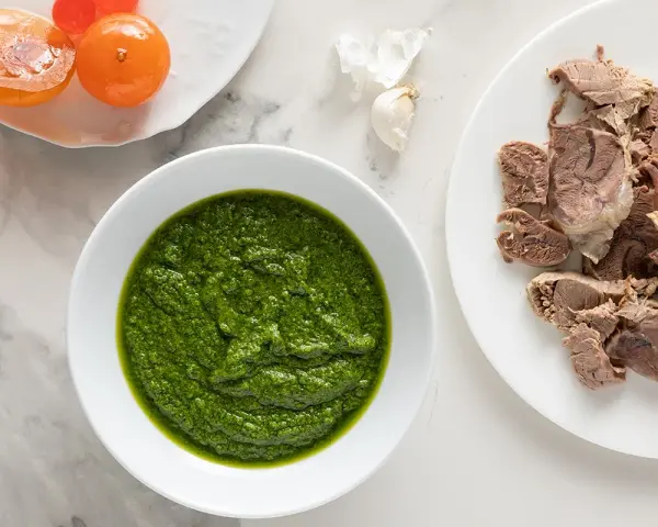 Una ciotola di vibrante salsa verde accanto a fette di carne bollita e pomodorini confit.