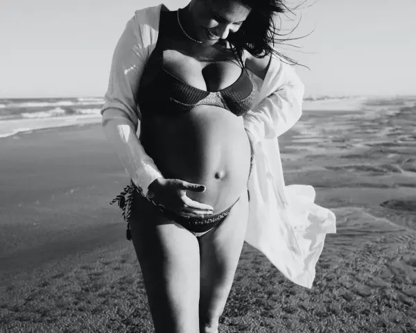 Donna incinta si gode il sole in spiaggia, accarezzando il pancione. La brezza marina le scompiglia i capelli.