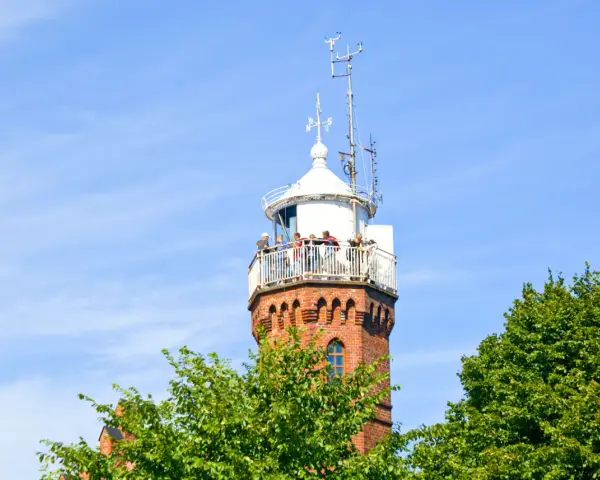 Widok na ceglaną latarnię morską w Ustce z tarasem widokowym, na którym stoją ludzie. Wokół zielone drzewa i błękitne niebo.