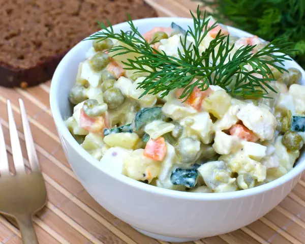 Ein Schüsselchen Olivier Salat, garniert mit Dill, neben einer Scheibe dunklem Brot und einer Gabel.