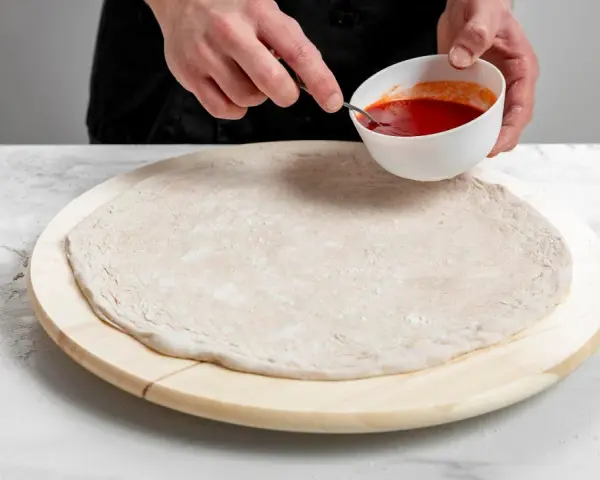 Przygotowanie ciasta na pizzę bez drożdży. Dłonie nabierają sos pomidorowy z miseczki, by rozprowadzić go na okrągłym spodzie.