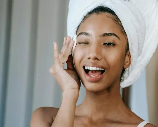 Une jeune femme avec une serviette sur la tête, appliquant un soin sur son visage. Elle semble heureuse, peut-être grâce aux bienfaits du collagène ou de l'acide hyaluronique.