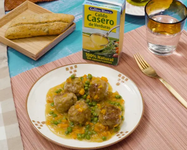Albondigas a la jardinera en un plato blanco, acompañadas de un pan rústico, un vaso de agua y un caldo de verduras.
