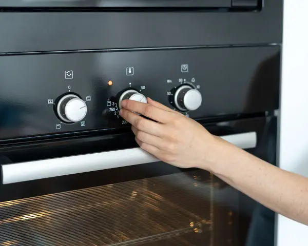 Hand dreht Temperaturregler am Ofen auf 250 Grad. Die Frage "wie lange Ofen vorheizen" wird durch das Einstellen der Temperatur beantwortet.