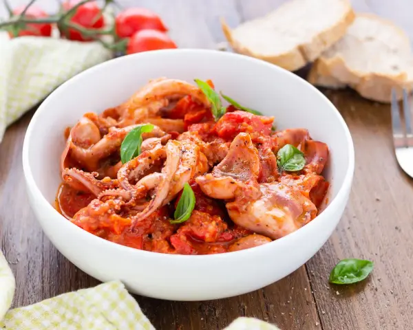 Totani in umido con pomodoro e basilico, serviti in una ciotola bianca, con pane e forchetta sullo sfondo.