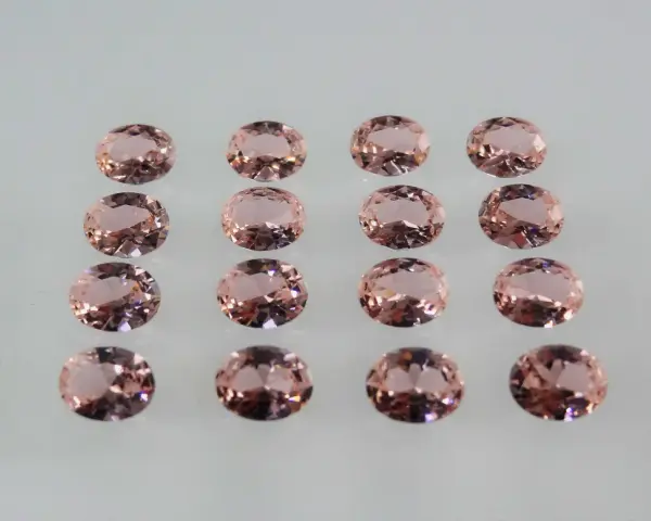 Sedici pietre di morganite ovali, rosa pesca, disposte in file ordinate. Le loro sfaccettature riflettono la luce, mostrando le belle morganite proprietà.