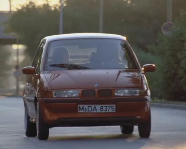 Pomarańczowy BMW E1 jedzie po drodze, światła odbijają się od karoserii.