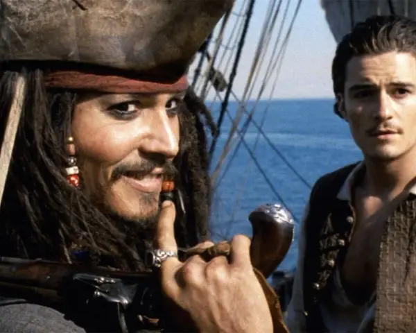 Jack Sparrow, z fajką i karabinem, uśmiecha się do Willa Turnera. Pewnie myśli o kolejnej butelce rumu.