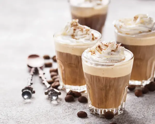 Tre bicchieri di deliziosa crema al caffè, guarniti con panna montata e cacao. Chicchi di caffè e cucchiaini decorati completano la scena.