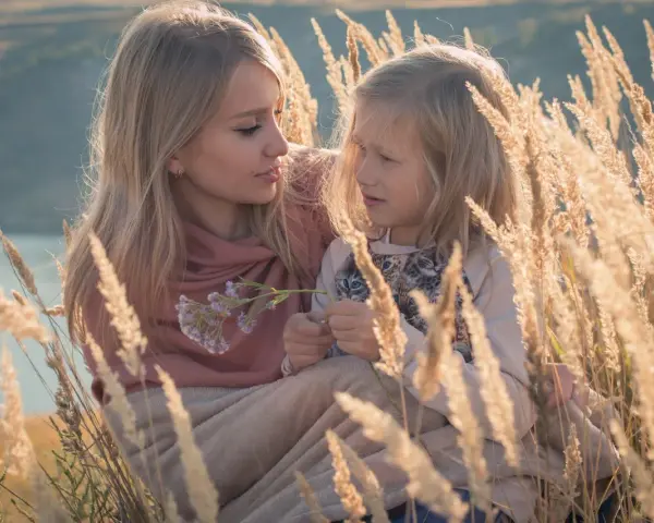 Matka i córka w trawach. Córka z autyzmem skupiona na kwiatku, matka z czułością patrzy na nią.