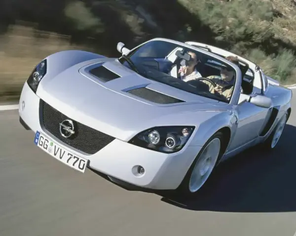 Srebrny Opel Speedster z dwoma pasażerami pędzi po krętej drodze.