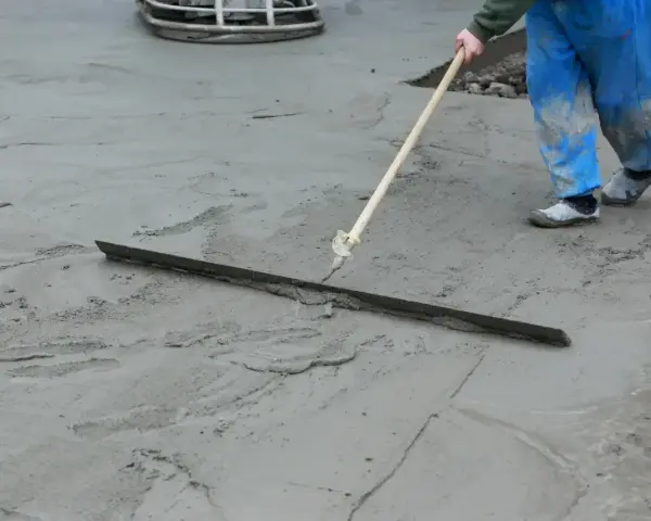 Pracownik w niebieskich ogrodniczkach wygładza świeży beton długą pacą. Trwa wygrzewanie posadzki.
