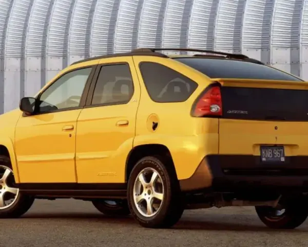 Żółty Pontiac Aztek z charakterystycznym tyłem i spojlerem, zaparkowany przed falistą, metalową ścianą.