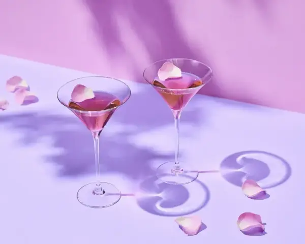 Elegancki Martini przepis: dwa kieliszki z różowym drinkiem, ozdobione płatkami róż, na fioletowym tle.