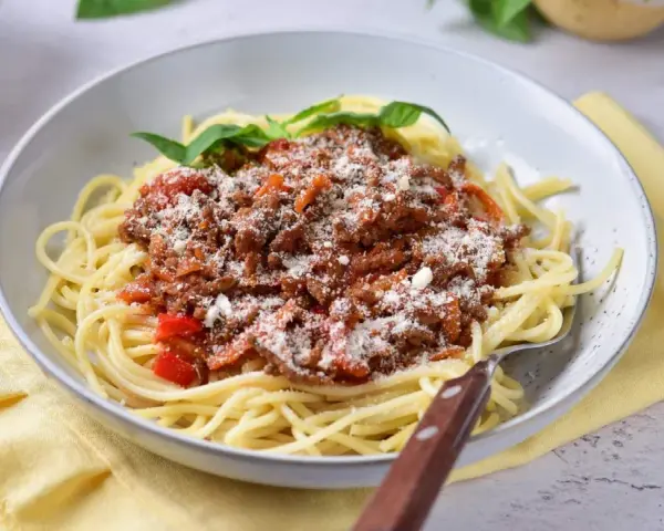 Pyszne spaghetti z bogatym sosem mięsnym na bazie passaty, posypane parmezanem i udekorowane listkami bazylii.