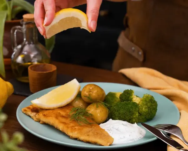 En hand pressar citron över en tallrik med panerad torsk, potatis, broccoli och en klick sås.