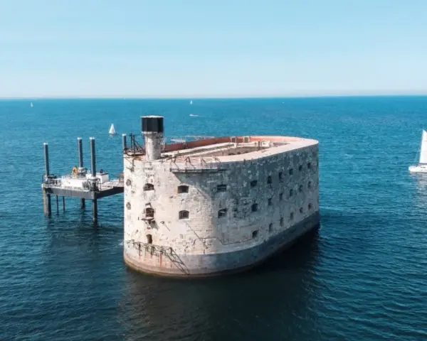 Fort Boyard, potężna twierdza na morzu, otoczona błękitną wodą. Obok platforma wiertnicza i żaglówka.
