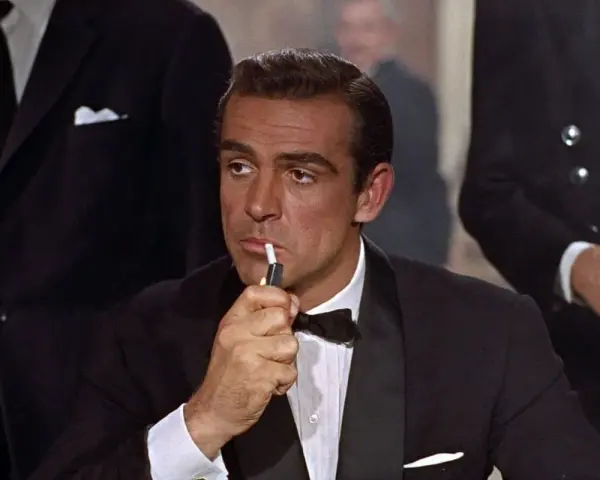 James Bond, elegante in smoking, accende una sigaretta con un'aria pensierosa.