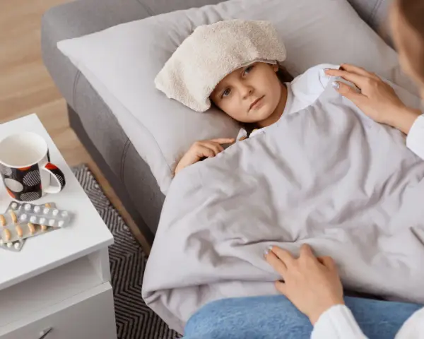 Chora dziewczynka z okładem na głowie leży pod kocem. Obok leki i kubek. Mama czuwa, zapewniając opiekę na dziecko.