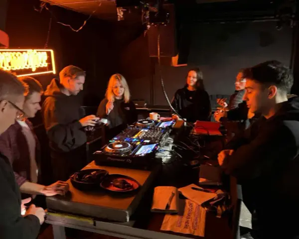 Grupa młodych ludzi przy konsoli DJ-skiej, uczących się, jak zostać DJ-em. Neonowy napis w tle.