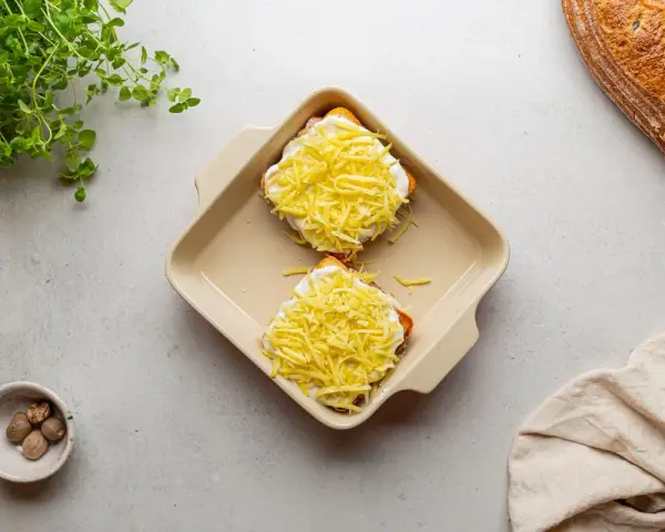 Dwa tosty z serem i beszamelem, przygotowane do zapieczenia jako croque madame. Obok gałka muszkatołowa i świeże zioła.