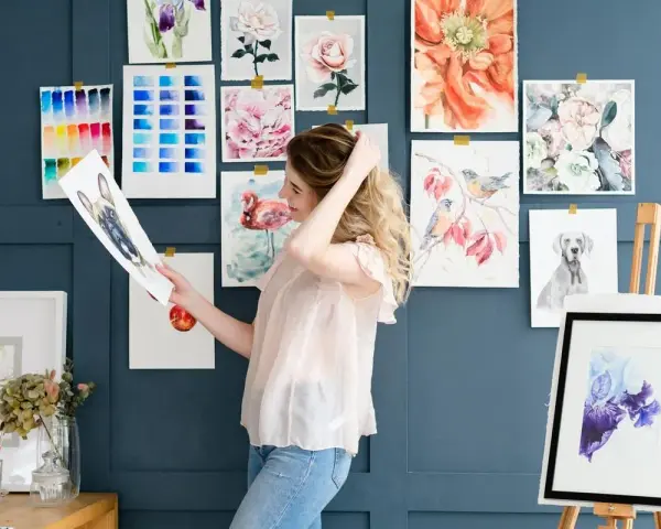 Mujer joven sonríe mientras revisa un dibujo de perro, rodeada de acuarelas de flores, aves y un flamenco. Un espacio inspirador para estudiar dibujo.
