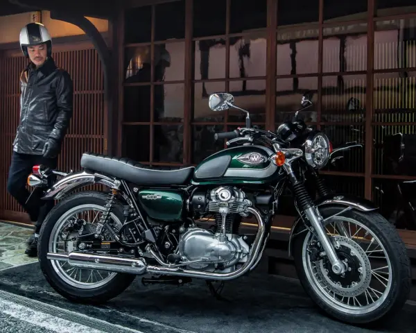 Mężczyzna w skórzanej kurtce i kasku stoi obok klasycznego motocykla Kawasaki W800 w kolorze ciemnozielonym.