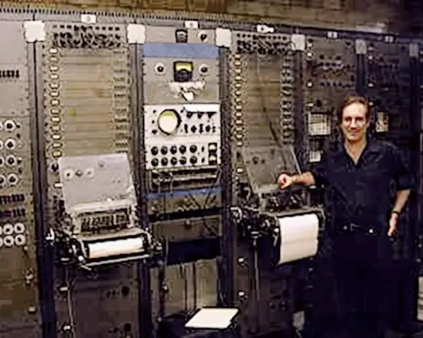 Mann steht neben einem riesigen, frühen Computer, dem ersten Synthesizer der Musikgeschichte.