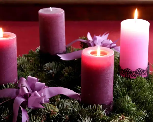 Drei brennende Kerzen auf einem Adventskranz aus Tannengrün mit lila Schleifen. Die dritte Kerze leuchtet hell.