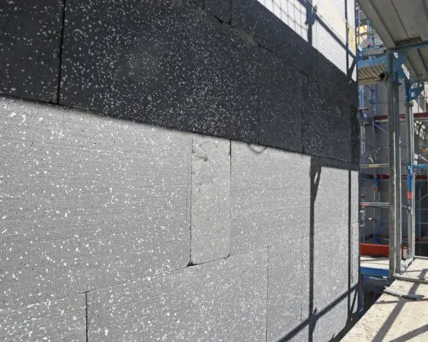 Gros plan sur des blocs de polystyrène gris, certains recouverts d'une couche noire, en cours d'installation pour une isolation par l'extérieur. Échafaudages visibles à droite.