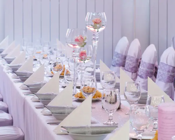 Vacker dukning med rosa rosor i glasvaser, vita servetter och silverfärgade skålar på en rosa bordsduk.