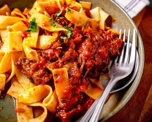 Breite Bandnudeln mit herzhaftem Ragu-Sauce, garniert mit frischer Petersilie. Ein Gabel liegt bereit.