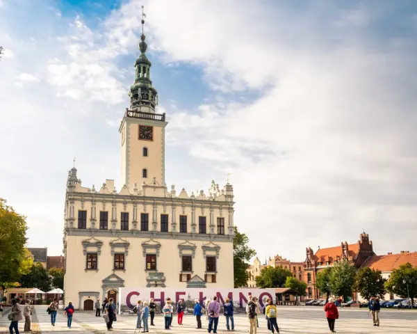 Rynek w Chełmnie z ratuszem i tablicą z napisem "Chełmno". Ludzie spacerują, podziwiając atrakcje miasta.