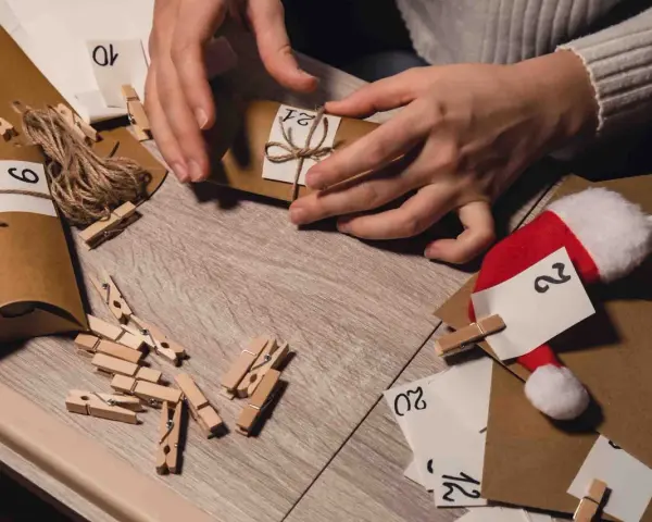 Händer som dekorerar en egen julkalender med nummer och snöre. Små klädnypor och en tomteluva syns också.