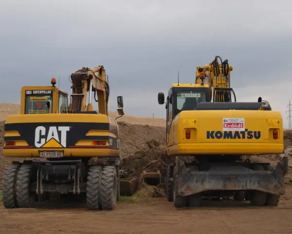 Dwie żółte koparki, CAT i Komatsu, stoją na placu budowy. Czy koparka ma dowód rejestracyjny? Na tablicy rejestracyjnej CAT widnieje KL 505 AE.