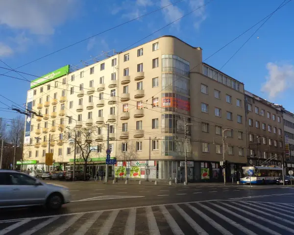Charakterystyczny budynek z balkonami i zaokrąglonym narożnikiem, przykład modernizmu w architekturze, na tle błękitnego nieba.