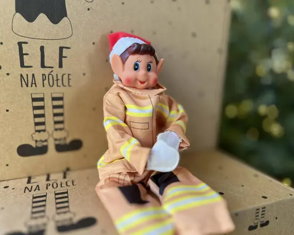 Uroczy elf na półce w stroju strażaka siedzi na kartonie z rysunkami elfich nóg.
