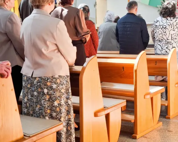 Ludzie stoją w kościele, czekając na dyspensę. Widać ławki i fragmenty ubiorów wiernych.