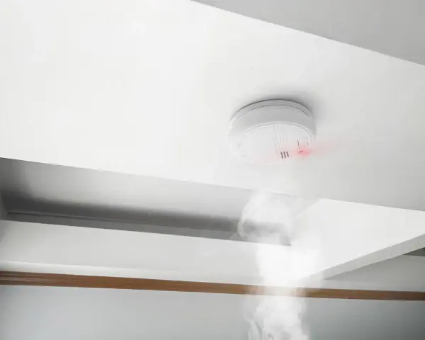 Detector de humo blanco en techo, emitiendo luz roja y humo. Cumpliendo la normativa de detectores de humo para seguridad.