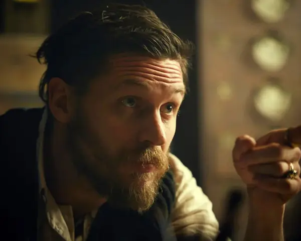 Mężczyzna z charakterystyczną fryzurą w stylu Peaky Blinders, z zarostem i pierścieniami na palcach, wskazuje palcem.