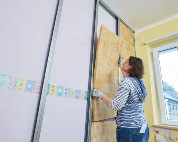 Frau baut eine Trockenbauwand, setzt Dämmmaterial in Metallständer ein.