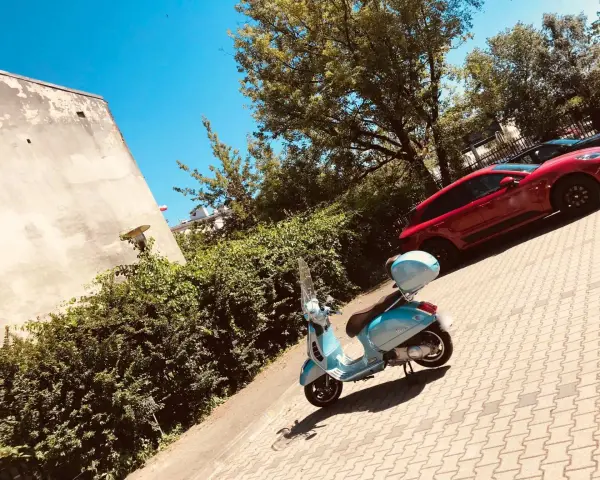 Błękitny skuter czeka na przejażdżkę. W tle czerwony samochód i zielone drzewa. Idealne miejsce na parking moto.