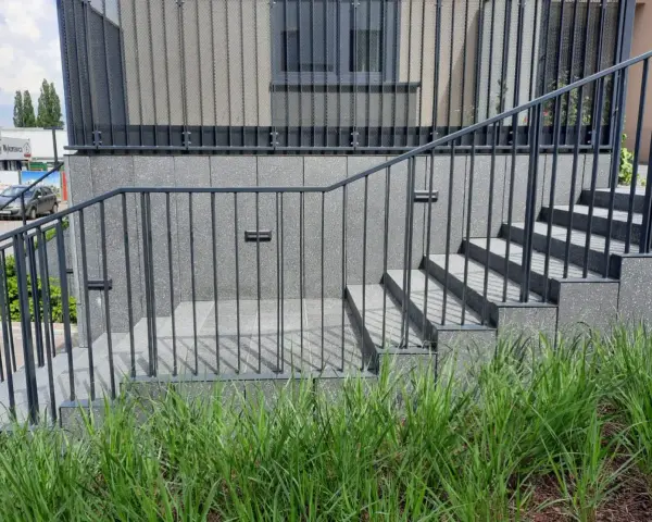 Nowoczesne schody zewnętrzne z metalową balustradą prowadzą do budynku. Wokół bujna zieleń.