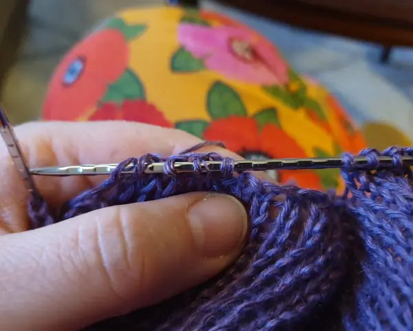 Närbild på en hand som stickar med lila garn. Nålen håller flera maskor, redo för nästa varv i blocka stickning.