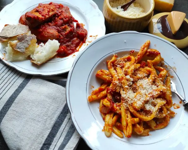 Pyszne maccheroni al ferretto w sosie pomidorowym, posypane parmezanem, obok talerz mięsa w sosie i chleba.
