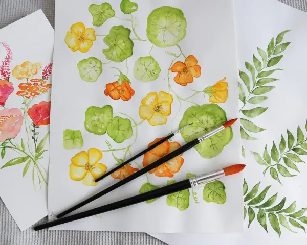 Flera akvarellmålningar med blommor och blad. En målning visar gula och orange blommor med gröna blad, en annan visar en bukett med rosa och orange blommor. Penslar ligger på pappret.